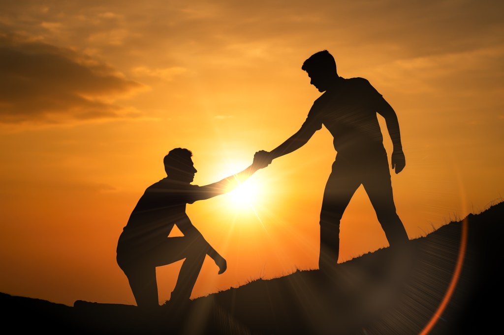kindness man reaching for another man to help up a hill sun in background